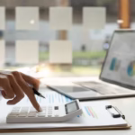 Osoba licząca koszty firmy przy biurku z laptopem i kalkulatorem – wirtualne biuro w Łodzi.